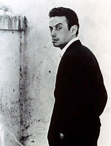 Lenny Bruce looking at the camera in a corner of a stone structure smoking a cigarette.